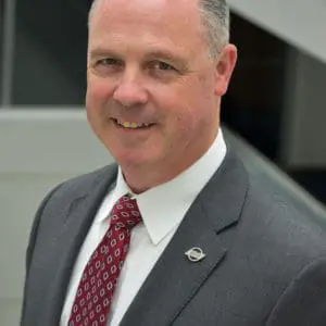 A man in a grey suit, white shirt, and red patterned tie smiles while standing in an indoor setting.