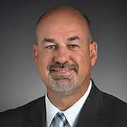 A bald man with a goatee is wearing a suit and tie, smiling against a dark background.