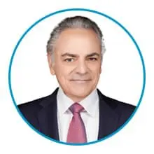 A person with gray hair is wearing a suit and a red tie, smiling and looking directly ahead, framed within a blue circle.