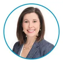 A woman with shoulder-length hair is smiling and wearing a blazer and a necklace, framed by a blue circular border.
