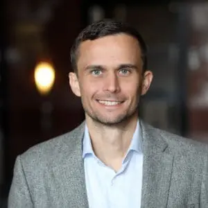 A man smiles while wearing a light blue shirt and a gray blazer.