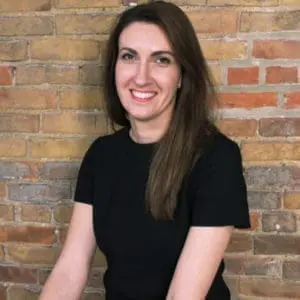 A person with long brown hair is smiling and standing in front of a brick wall.