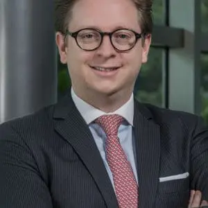 A person with glasses and a suit stands with arms crossed, smiling in front of a window.