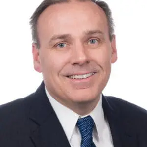 A man dressed in a dark suit, white shirt, and blue tie is smiling against a white background.
