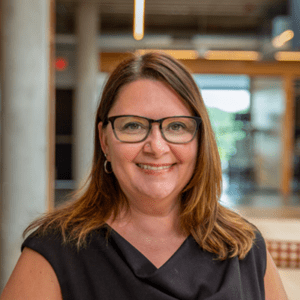 A woman with glasses and shoulder-length brown hair is smiling while wearing a black shirt, standing indoors with contemporary architecture in the background.