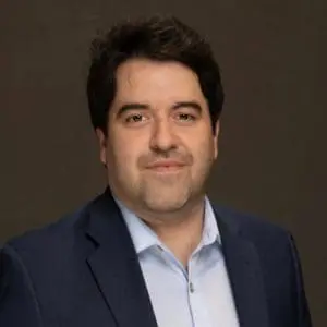 A person with dark hair is wearing a dark blue suit and light blue shirt against a plain background.