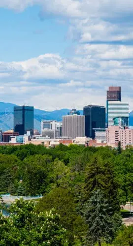 A vibrant city skyline rises above a lush, green urban park with mountains in the background under a partly cloudy sky.