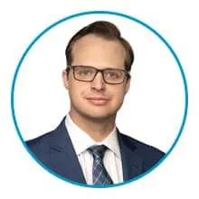 A person wearing glasses and a suit with a tie is shown inside a circular frame.