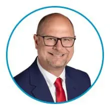 A smiling man wearing glasses, a blue suit, and a red tie is within a blue circular border.