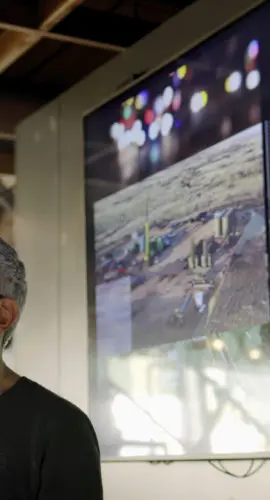 Stock photograph of a man conducting a seminar / lecture with the aid of a large screen. The screen is displaying data & designs concerning low carbon electricity production with solar panels & wind turbines. These are juxtaposed with images of conventional fossil fuel oil production.