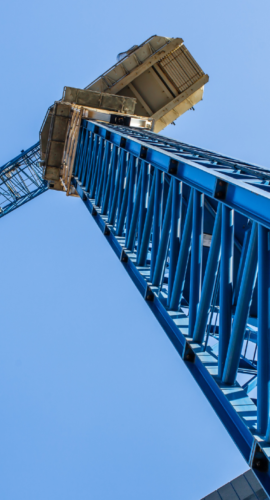 Upward view of a blue construction crane against a clear sky with modern skyscrapers in the background, representing commercial real estate finance, lending transactions, and the complex carveout provisions and negotiation strategies that protect lenders and borrowers in major real estate deals.