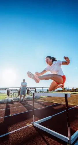 A female collegiate heptathlete powers over a hurdle at full speed during an intense solo training session with her coach observing nearby.