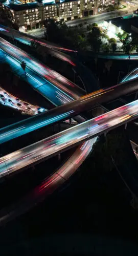 Aerial long exposure photograph of a multi-level freeway interchange near Downtown Los Angeles at night, with light trails from vehicles streaking red and white across elevated lanes and on and off ramps. Residential and commercial buildings line the upper edge of the frame, illuminated against the dark cityscape. Palm trees are visible along the median strips between the carriageways.