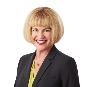 A smiling woman with blonde hair, wearing a black blazer over a yellow blouse, poses against a white background.