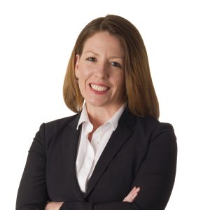 A person with long hair wearing a white shirt and black blazer is smiling against a plain background.