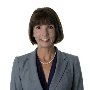 A professional portrait of a woman with short hair wearing a gray blazer, a blue blouse, and a pearl necklace.