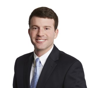 A smiling individual wearing a dark suit, light blue striped tie, and a white shirt against a plain background.