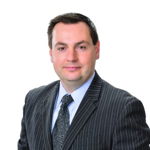 A person wearing a professional suit and tie poses against a plain background.