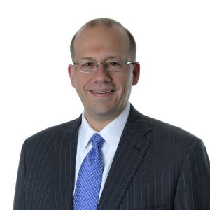 A smiling person wearing glasses, a blue patterned tie, and a dark pinstriped suit poses against a plain background.