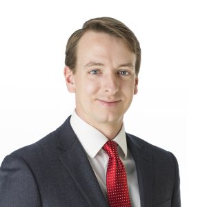 A man wearing a suit with a red tie is smiling at the camera against a white background.