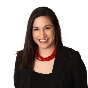 A smiling person with dark hair wearing a black blazer and a red beaded necklace.