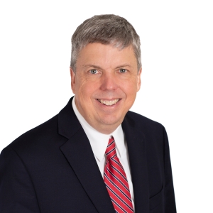 A smiling individual with short hair wearing a navy blazer and a red striped tie against a white background.