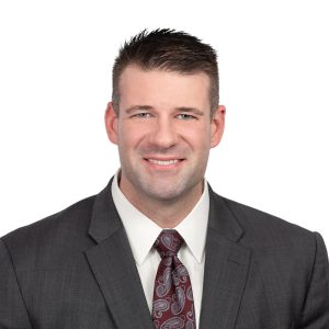 A smiling man wearing a dark suit with a red patterned tie and a white shirt poses for a professional portrait.