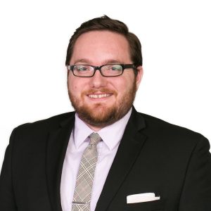 A smiling individual wearing glasses, a black suit jacket, a light purple shirt, and a patterned tie.