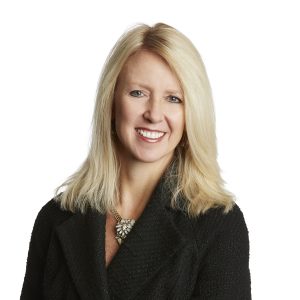 A smiling woman with blonde hair is wearing a black jacket and a decorative necklace.