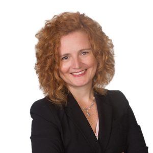 A smiling woman with curly hair is wearing a professional black jacket over a pink shirt against a neutral background.