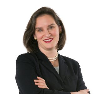 A smiling woman with short dark hair, wearing a black blazer, white blouse, and a pearl necklace, is posing with her arms crossed.