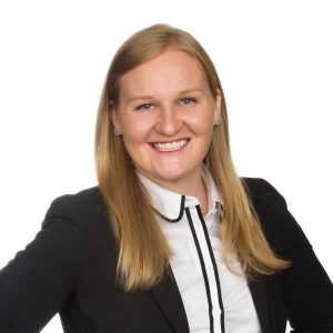 A smiling individual with long hair is dressed in professional attire featuring a black blazer and a white collared shirt with black piping.