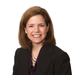 A smiling individual with shoulder-length hair wearing a black blazer and a purple blouse.