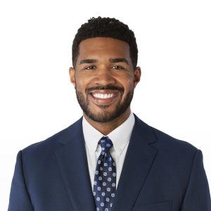 A person with a neatly trimmed beard is smiling and wearing a blue suit with a polka dot tie.