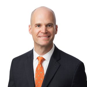 A smiling individual with a shaved head, wearing a dark suit with a white shirt and an orange tie.