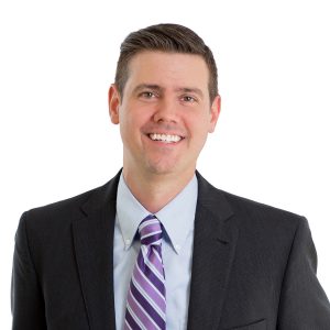 A smiling individual wearing a suit with a striped purple tie against a white background.