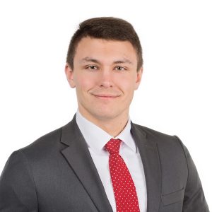 A smiling individual wearing a gray suit with a red tie against a white background.