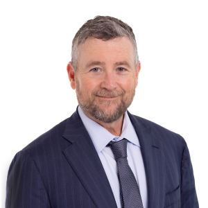 A bespectacled person with a beard is wearing a suit and tie against a plain background.