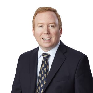 A person wearing a dark suit, white shirt, and patterned tie is posing against a white background.