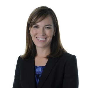 A smiling woman wearing a black blazer over a blue blouse against a white background.