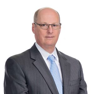 A man wearing a suit and tie stands against a plain background.