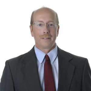 A person with a neutral expression is wearing a gray suit, white shirt, and a red tie against a white background.
