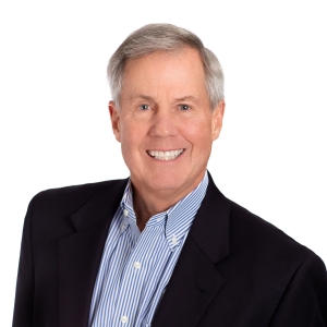 A smiling individual wearing a black blazer over a blue-striped shirt against a white background.