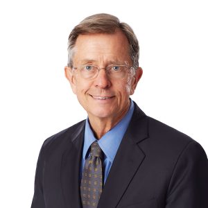 A smiling person wearing glasses, a dark suit, a blue shirt, and a patterned tie against a white background.