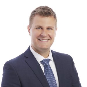 A smiling individual wearing a blue suit and tie against a white background.