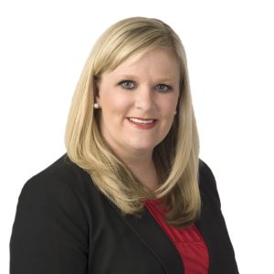 A smiling woman with blonde hair wearing a black jacket over a red top against a white background.
