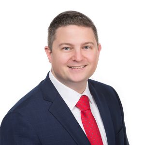 A smiling individual wearing a suit with a red tie poses against a white background.