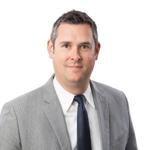 A man in a gray suit with a blue tie is smiling at the camera against a white background.