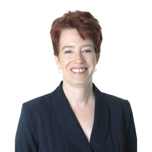 A smiling person with short red hair wearing a dark blazer over a light-colored top against a white background.