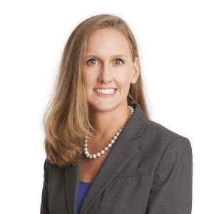 A smiling person with blond hair wearing a pearl necklace and a gray blazer over a blue top.